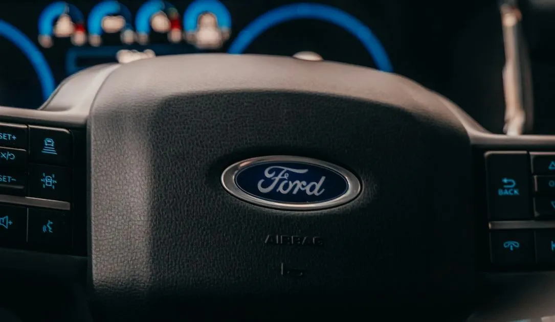 Volante de un coche Ford con el logotipo de la marca en el centro. El tablero de instrumentos con luces azules es visible al fondo, junto con algunos controles del vehículo en el lado izquierdo del volante.